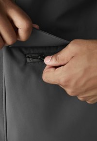 Black, smooth fabric pocket being opened, featuring a concealed zipper with a pull tab. Hands gripping the edge of the pocket.