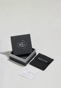 Schwarze Schmuckschatulle mit einem weißen Logo, geöffnet, um eine graue Innenschachtel, einen schwarzen Beutel und ein weißes Etikett mit goldener Schrift zu zeigen.