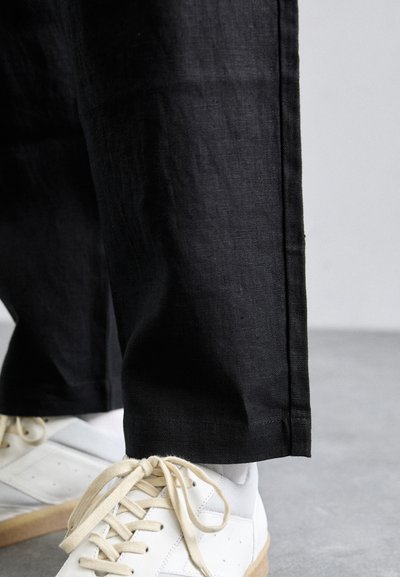 Spodní část nohou v černých rovnějších kalhotách a bílých teniskách s béžovými tkaničkami na hladké šedé podlaze.