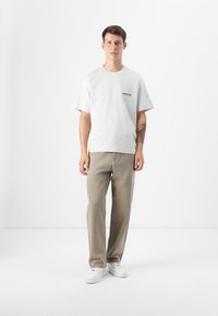 Un joven está de pie contra un fondo blanco, vistiendo una camiseta gris clara con un logo, pantalones beiges y zapatillas blancas. Tiene el pelo corto de color castaño.