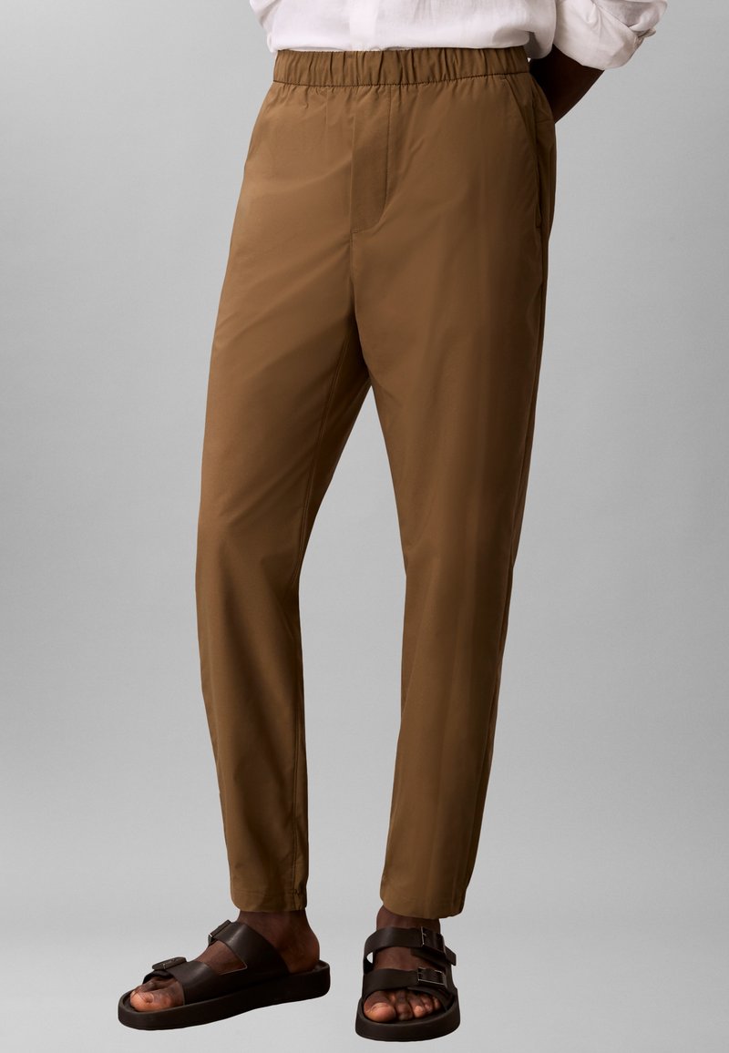 Pantalon à taille élastique marron porté avec une chemise blanche, accompagné de sandales noires à double bride sur une personne devant un fond gris.
