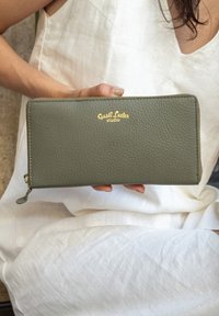 Portafoglio in pelle verde con finitura testurizzata, chiusura a zip e logo in oro "Gusti Leder studio". Tenuto a mano su uno sfondo chiaro.