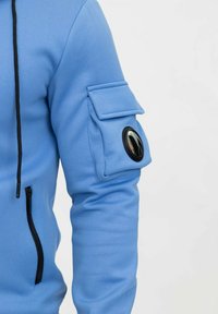 Sweat à capuche bleu avec une texture lisse, doté d'une poche zippée, d'un patch sur la manche et d'un accent circulaire. Cordon de serrage noir inclus.