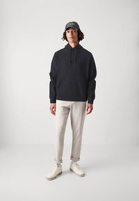 Fekete kapucnis pulóver húzózsinórral, világosszürke, szűkített nadrág, krémszínű sportcipő, kiegészítve egy sötét sapkával, logó részlettel.