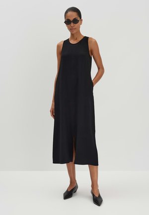 Schwarzes ärmelloses Maxikleid mit Seitentaschen und einem vorderen Schlitz, aus leichtem Stoff. Kombiniert mit flachen, spitz zulaufenden schwarzen Schuhen und Sonnenbrille.