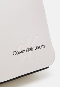 Bílá taška z umělé kůže s raženým logem "CK" a černým potiskem "Calvin Klein Jeans", s hladkým povrchem a zakřiveným tvarem.