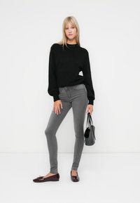 Pull en tricot noir, jeans skinny gris et ballerines bordeaux. Le mannequin tient un petit sac à main noir. Design simple et épuré.