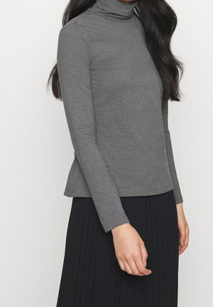 Vrouw die een grijs langharig coltrui-top draagt, gecombineerd met een zwarte geplooide rok tegen een effen witte achtergrond.