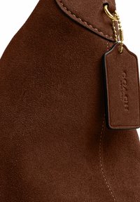 Cuir suédé marron avec des coutures détaillées et une étiquette en cuir attachée par une chaîne à billes dorée étiquetée "Coach New York."