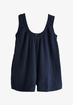 Kvinde iført en mørkeblå ærmeløs top og en rund stenperle halskæde, der rører ved sit hår med én hånd prydet med armbånd.