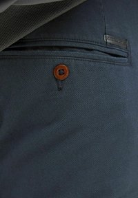 Donkerblauwe stof met een gestructureerde weving, voorzien van een bruine knop en een gestikte zakdetail. Inclusief een klein merketiket.