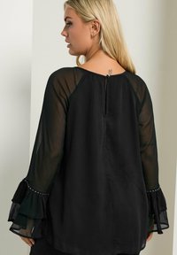 Blusa nera con maniche lunghe trasparenti, polsini arricciati e chiusura sul retro con bottoni. Il tessuto ha una consistenza morbida con leggeri dettagli arricciati.