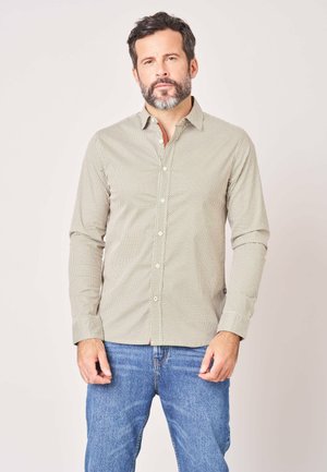 Man met donker haar en baard, die een licht patroon overhemd met knopen en blauwe spijkerbroek draagt, staat tegen een effen achtergrond.