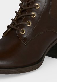 Gros plan d’une botte cheville en cuir marron avec des lacets ronds passés à travers des œillets dorés et un talon bas empilé.