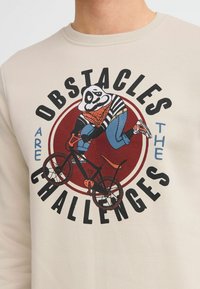 Beige sweatshirt featuring a graphic of a panda in a striped shirt riding a black bicycle, with the text "Obstacles are the Challenges."