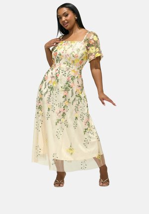 Frau posiert in einem cremefarbenen Midikleid mit transparenter floraler Stickerei, eckigem Ausschnitt und flatternden Ärmeln, trägt goldene Riemchen-Highheels.