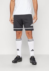 Čierne športové šortky so bielymi pruhmi a logom Adidas, v kombinácii s bielymi ponožkami s čiernymi pruhmi a čiernymi kopačkami.