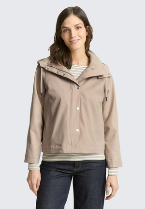 Frau trägt eine beige Kapuzenjacke mit Druckknöpfen über einem gestreiften Langarmshirt und dunklen Jeans, steht vor einem schlichten hellen Hintergrund.