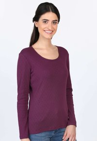 Langes lila Hemd mit Rundhalsausschnitt, gerippter Textur und subtilem geometrischen Muster. Getragen mit blauen Jeans.