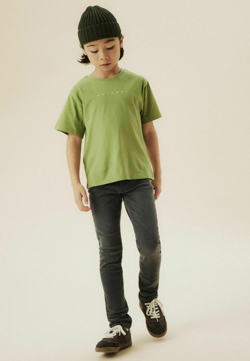 T-shirt vert à manches courtes avec un col rond, associé à un jean foncé. La tenue est complétée par un bonnet en tricot foncé et des chaussures décontractées.