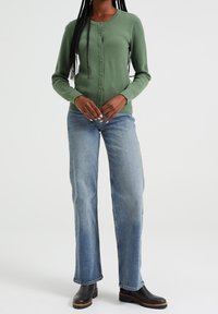 Green textured knit cardigan with round neckline and button front, paired with light blue straight-leg jeans and black ankle boots.