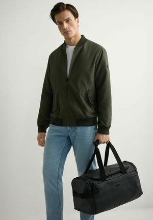 Młody mężczyzna w ciemnozielonej kurtce typu bomber i niebieskich dżinsach, trzymający czarną torbę sportową za uchwyty na jednolitym jasnym tle.