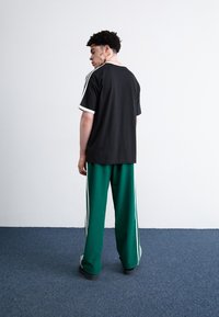 Črna majica s belimi obrobami, v kombinaciji z zelenimi hlačami, ki imajo bele stranske črte. Model je obrnjen od zadaj pred nevtralno ozadje.