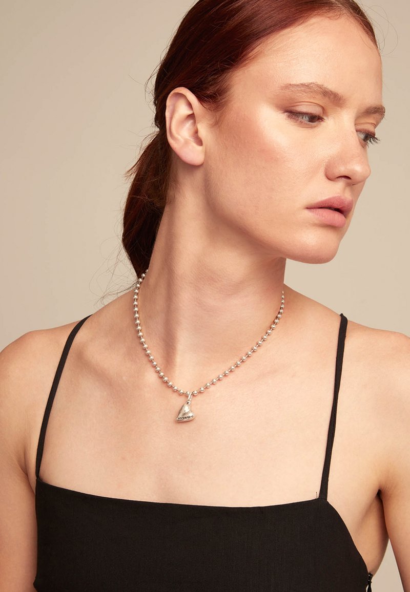 Collar de cuentas de plata con un colgante triangular, llevado sobre la piel desnuda. Modelo con una blusa de tirantes negros, fondo neutro.