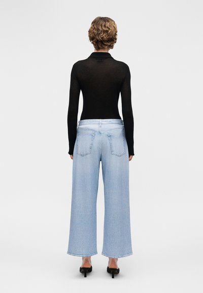 Άτομο με κοντά κυματιστά μαλλιά που φοράει μαύρο μακρυμάνικο μπλουζάκι, ανοιχτό μπλε παντελόνι φαρδιάς γραμμής και μαύρα παπούτσια με τακούνι, όρθιο με την πλάτη προς το μέρος μας.