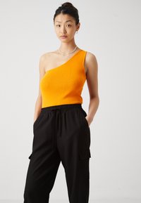 Naine, kes kannab varrukateta oranži üheõlalist topi ja lõdvaid musti pükse, käed taskutes, seisab ühevärvilise tausta ees.