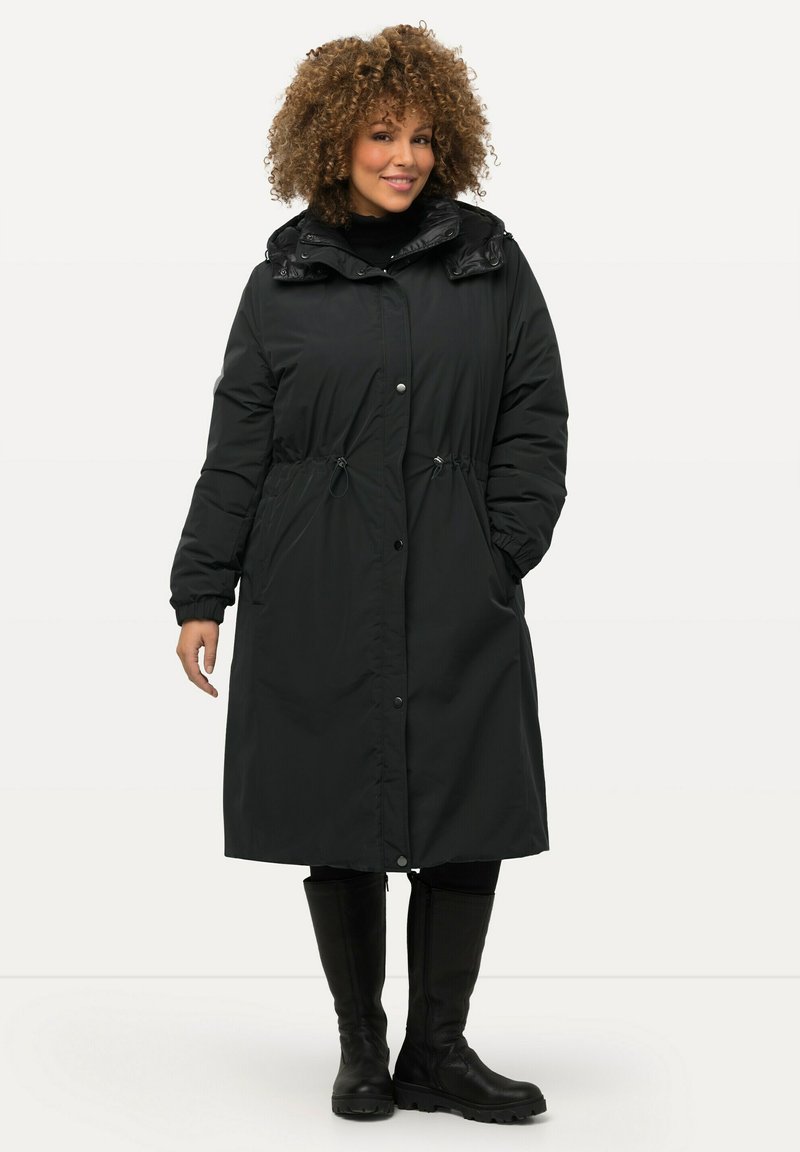 Mujer con cabello rizado que lleva una larga chaqueta acolchada negra con capucha y cordones, y botas negras hasta la rodilla, de pie y sonriendo.