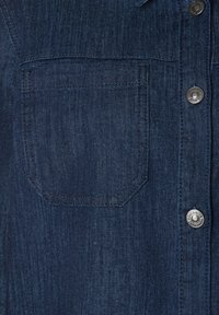 Dunkelblaues Denimhemd mit einer Brusttasche und metallenen Druckknöpfen, die vertikal auf der rechten Seite angeordnet sind.