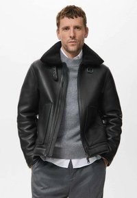 Chaqueta de cuero negra con un cuello de piel sintética desmontable, cierre de cremallera y bolsillos laterales, llevada sobre un suéter gris y una camisa blanca.