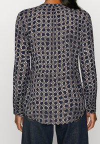 Långärmad blus i mörkgrått med ett cirkulärt mönster. Har rund halsringning och en knappdetalj på baksidan. Slät tygtextur.