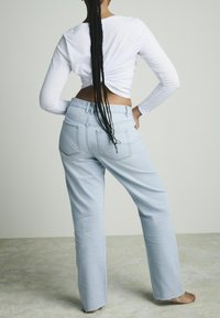 Lichtblauwe denim broek met een ontspannen pasvorm, ruwe zoom en twee achterzakken. Gepaard met een witte langslavende shirt, bekeken van achteren.