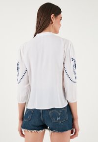 Blouse blanche avec de longues manches bouffantes, ornée de motifs brodés bleus sur les épaules. Portée avec un short en denim, texture lisse.