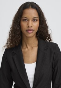 Blazer noir à coupe ajustée, avec des revers crantés, associé à un haut blanc à col rond. Le tissu a une texture lisse.