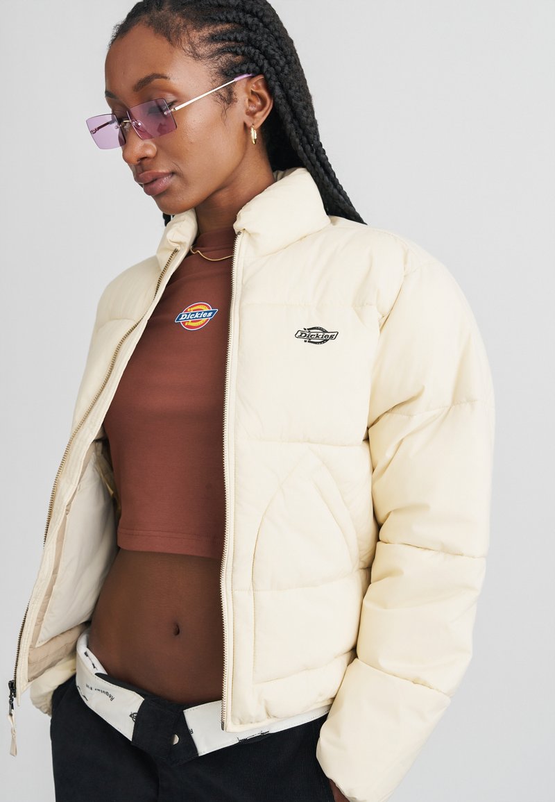 Giacca imbottita di colore crema, con chiusura zip, patch con logo e design trapuntato. Indossata sopra un top corto marrone, con pantaloni in velluto a coste neri.