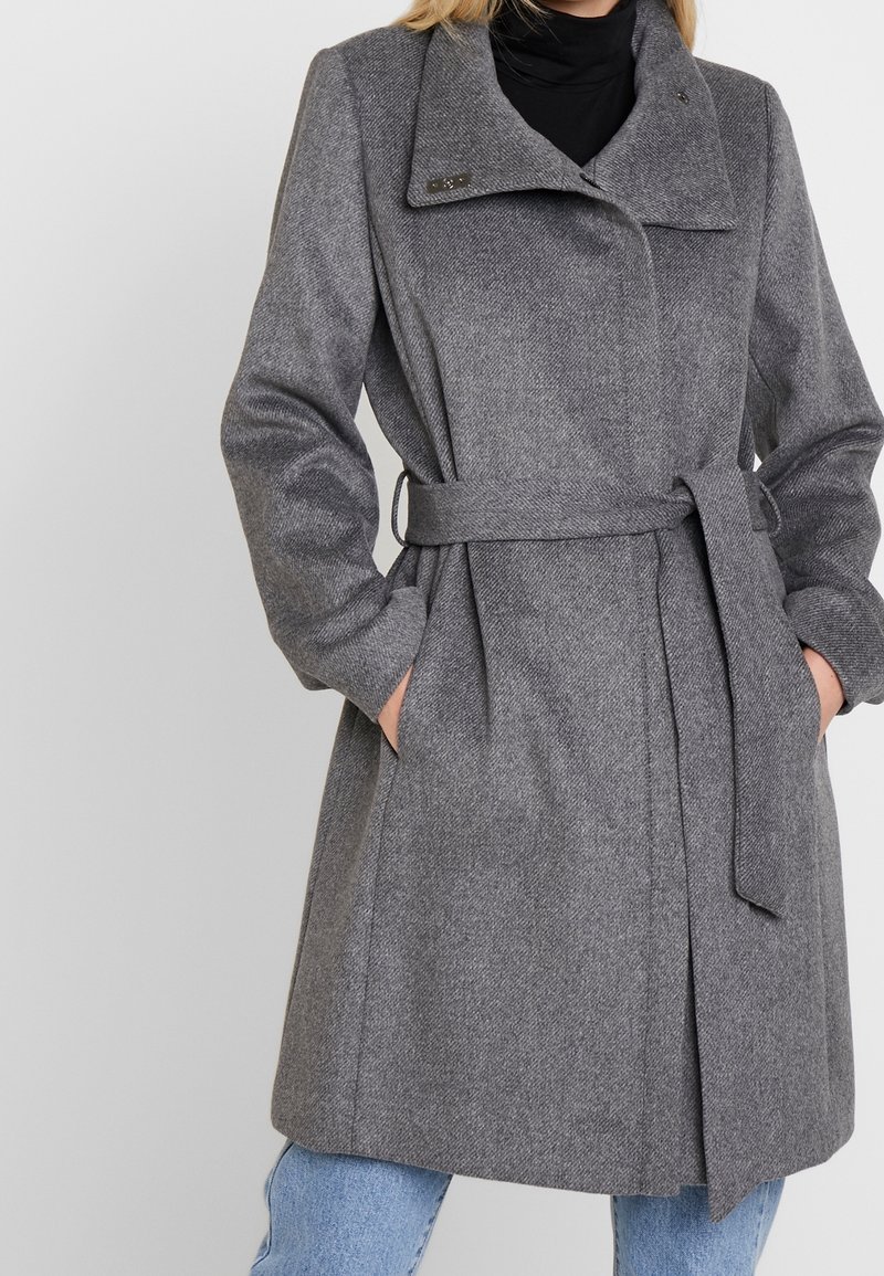 Cappotto grigio a spina di pesce con ampio colletto, vita cintrata e tasche a toppa. Il tessuto ha una finitura strutturata e presenta una silhouette sartoriale.