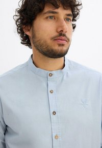 Camicia azzurra chiara con colletto e bottoni in legno, con un emblema ricamato di un cervo sul lato sinistro del petto. Tessuto di cotone morbido.