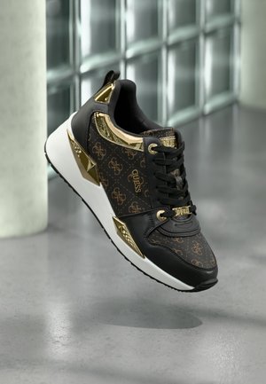 Czarny i złoty sneaker Guess z monogramem, białą podeszwą i metalicznymi złotymi akcentami, zawieszony nad szarą podłogą z tłem w postaci ściany z bloków szklanych.