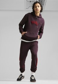 Burgunder Kapuzenpullover mit rotem besticktem Logo, kombiniert mit passenden Hosen, die rote Seitestreifen haben. Stilisiert mit schwarzen Sneakern und weißen Socken.