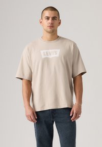 Beige bomulls t-skjorte med brede korte ermer og en hvit Levi's-logo på brystet. Kombinert med mørkeblå denimjeans.