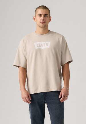 Beige Baumwoll-T-Shirt mit breiten, kurzen Ärmeln und einem weißen Levi's-Logo auf der Brust. Kombiniert mit dunkelblauen Jeans.