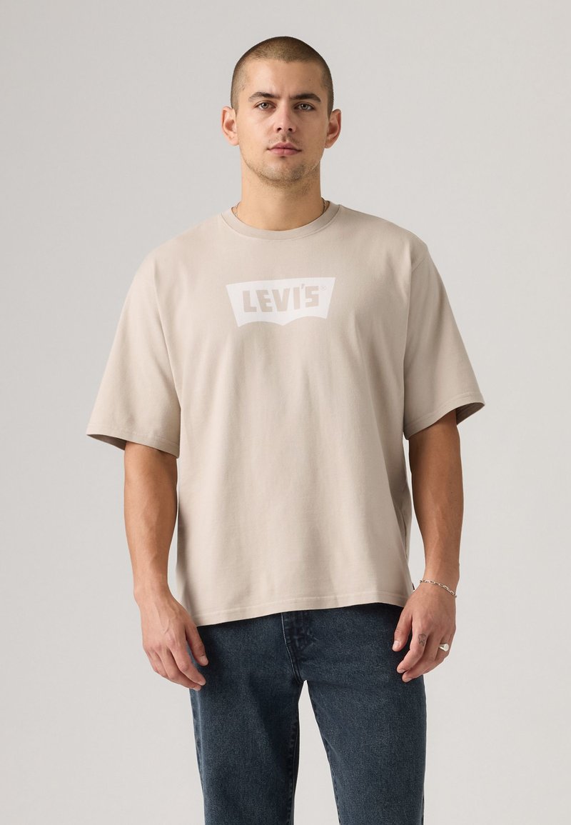 Beige bomulls t-skjorte med brede korte ermer og en hvit Levi's-logo på brystet. Kombinert med mørkeblå denimjeans.