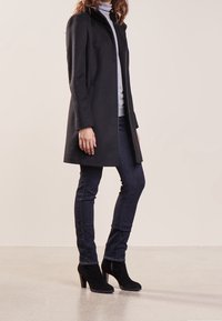 Schwarzer Wollmantel mit hohem Kragen, Knopfverschluss und geradem Schnitt, kombiniert mit dunkelblauen Jeans und schwarzen Wildleder-High-Heel-Stiefeletten.