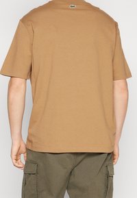 Brun t-shirt med kort ärm i bomull. Har rund hals och en liten grön logotyp på övre delen av ryggen. Slät struktur, avslappnad passform.