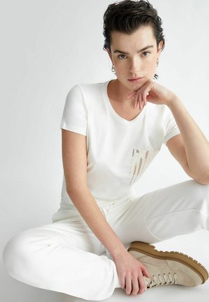 T-shirt bianca a maniche corte con un dettaglio decorativo a striscia verticale dorata sul lato sinistro. Indossata con pantaloni bianchi e stivali beige con lacci.