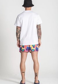 Camiseta blanca de algodón y pantalones cortos de colores con estampado de osos en rosa, verde, azul y amarillo. Sombrero tipo bucket negro y sandalias azules.