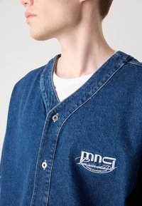 Denimjakke i mellomblått med knappelukking, v-hals og brodert logo på brystet. Har en teksturert stofffinish.